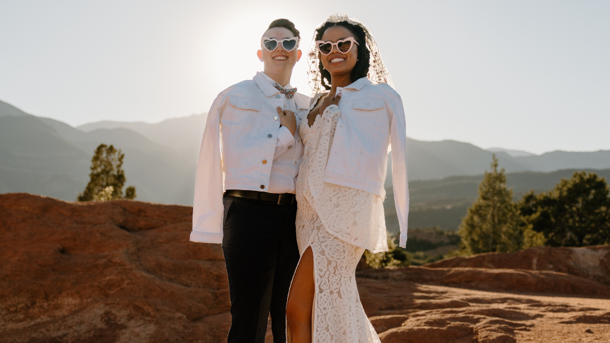A same-sex bridal couple poses together during a modern outdoor elopement at sunset in a desert mountain landscape with red rock formations. One bride wears dark dress pants, a white button-down shirt, and a bow tie, while the other wears a lace wedding dress with a high slit and a short veil. Both brides are smiling and wearing matching white, heart-shaped sunglasses with matching white denim jackets draped over their shoulders.