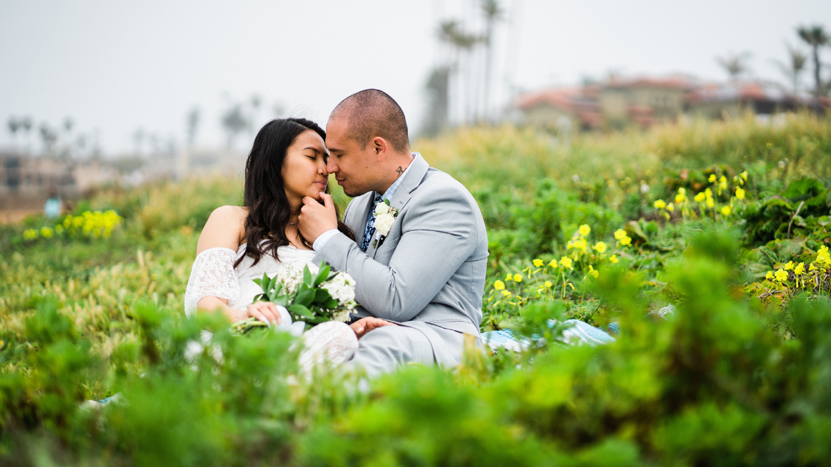 A bride and groom share a tender moment sitting together on a blue patterned blanket in a lush, green coastal field filled with small yellow wildflowers. The groom, wearing a light gray suit with a floral tie and white boutonniere, gently touches the chin of the bride, who is wearing an off-the-shoulder lace wedding dress, holding a white flower bouquet, and resting her eyes with a peaceful smile. The foggy background shows faint silhouettes of palm trees and coastal buildings.