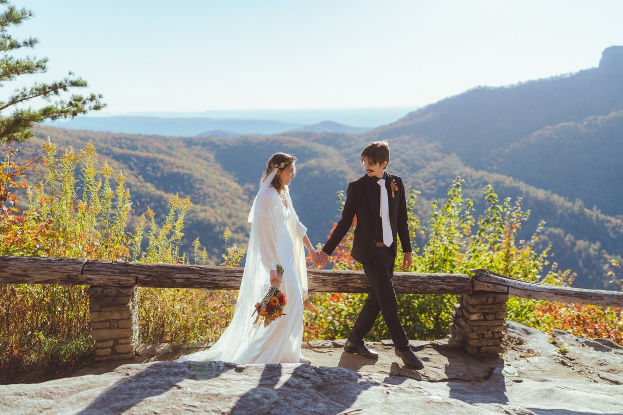 Bride and groom holding hands at a mountain overlook in North Carolina with autumn colors and Blue Ridge Mountain views.
