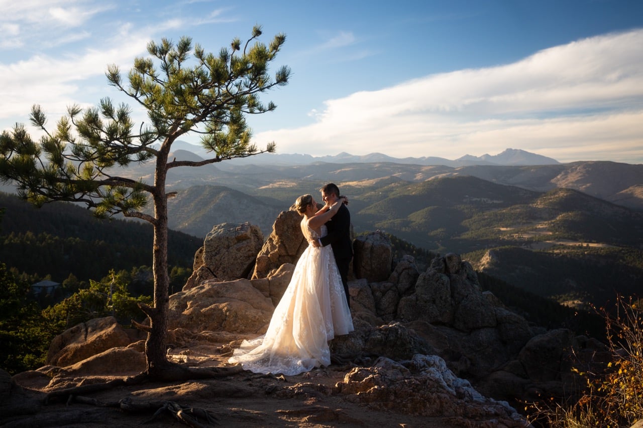 Bride and groom embracing on a rocky mountain overlook in Colorado at sunset with a lone pine tree and sweeping mountain views.