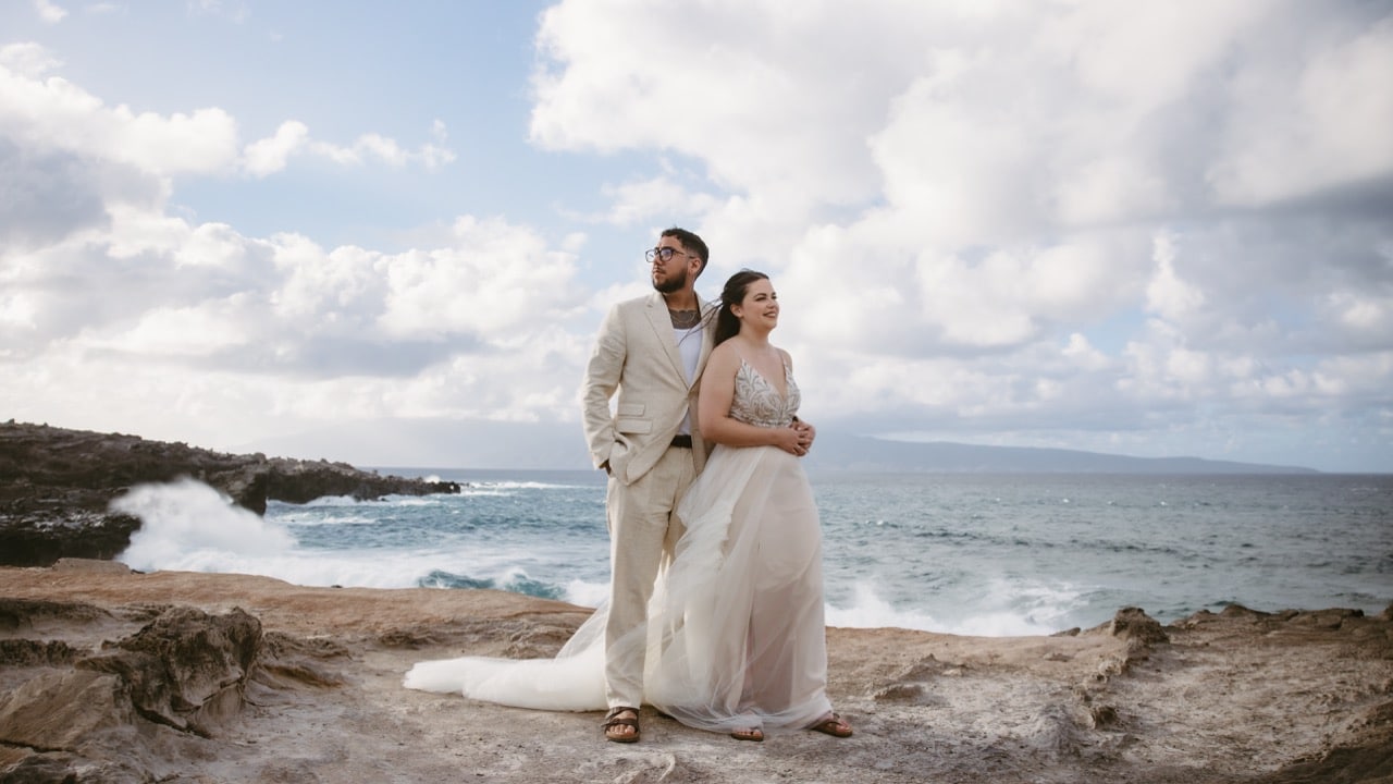Bride and groom standing on a rocky Maui coastline with ocean waves and dramatic clouds in the background, wearing light neutral wedding attire.