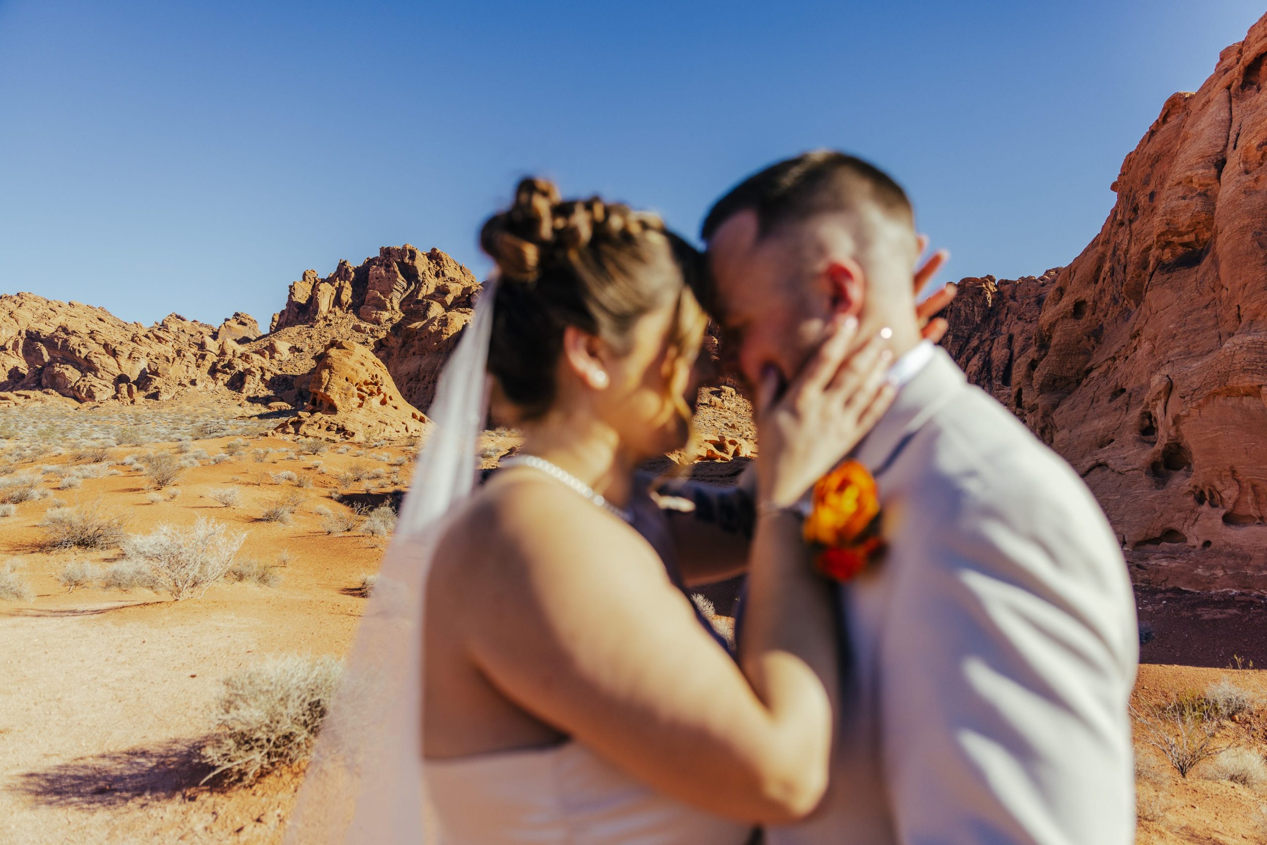 Bride and groom share an intimate moment during their desert elopement, framed by red rock formations and a clear blue sky, with the bride touching the groom’s face as they lean in together.