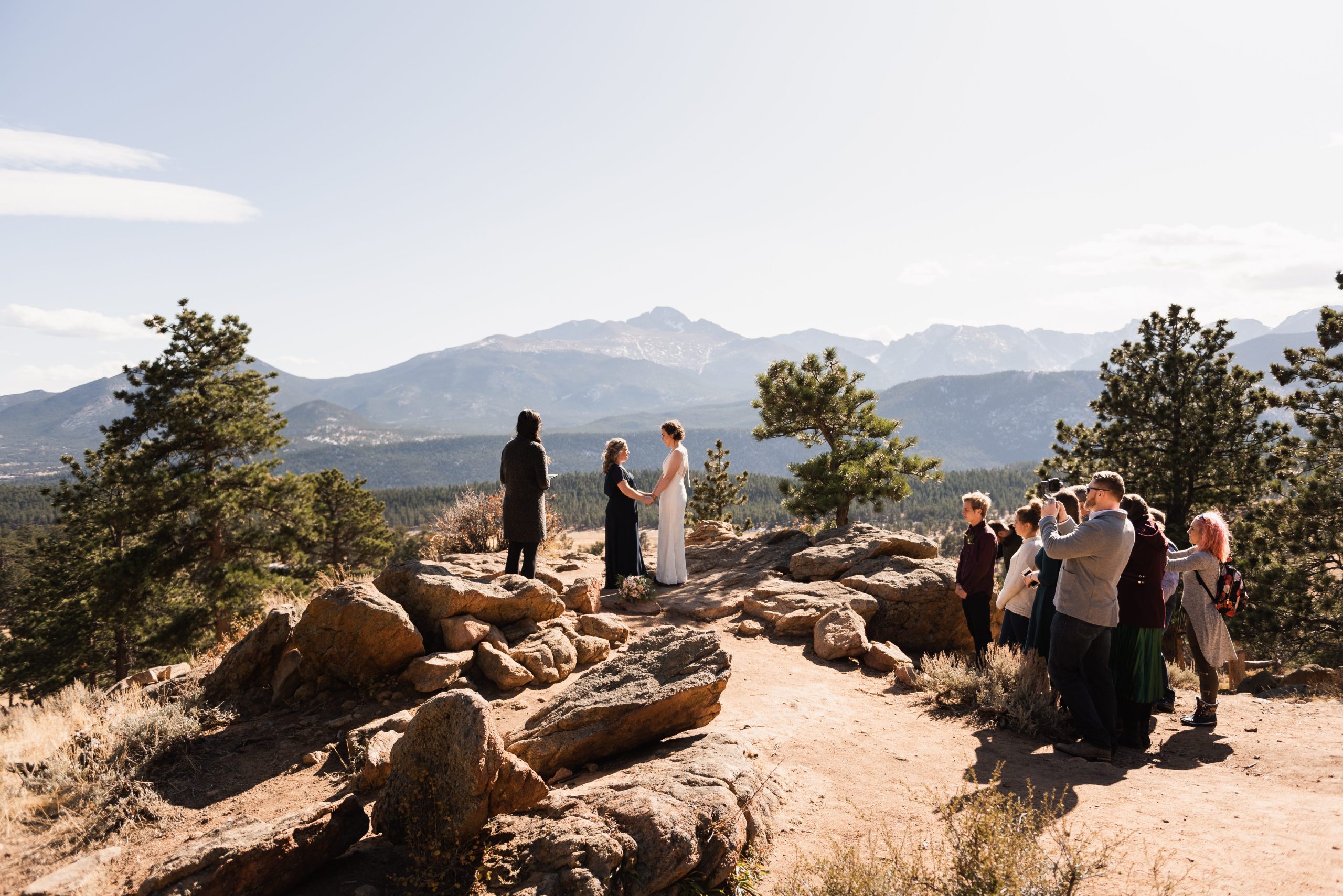 Couple exchange vows during a small mountaintop wedding ceremony with a handful of guests, standing on a rocky overlook surrounded by pine trees and mountain peaks.