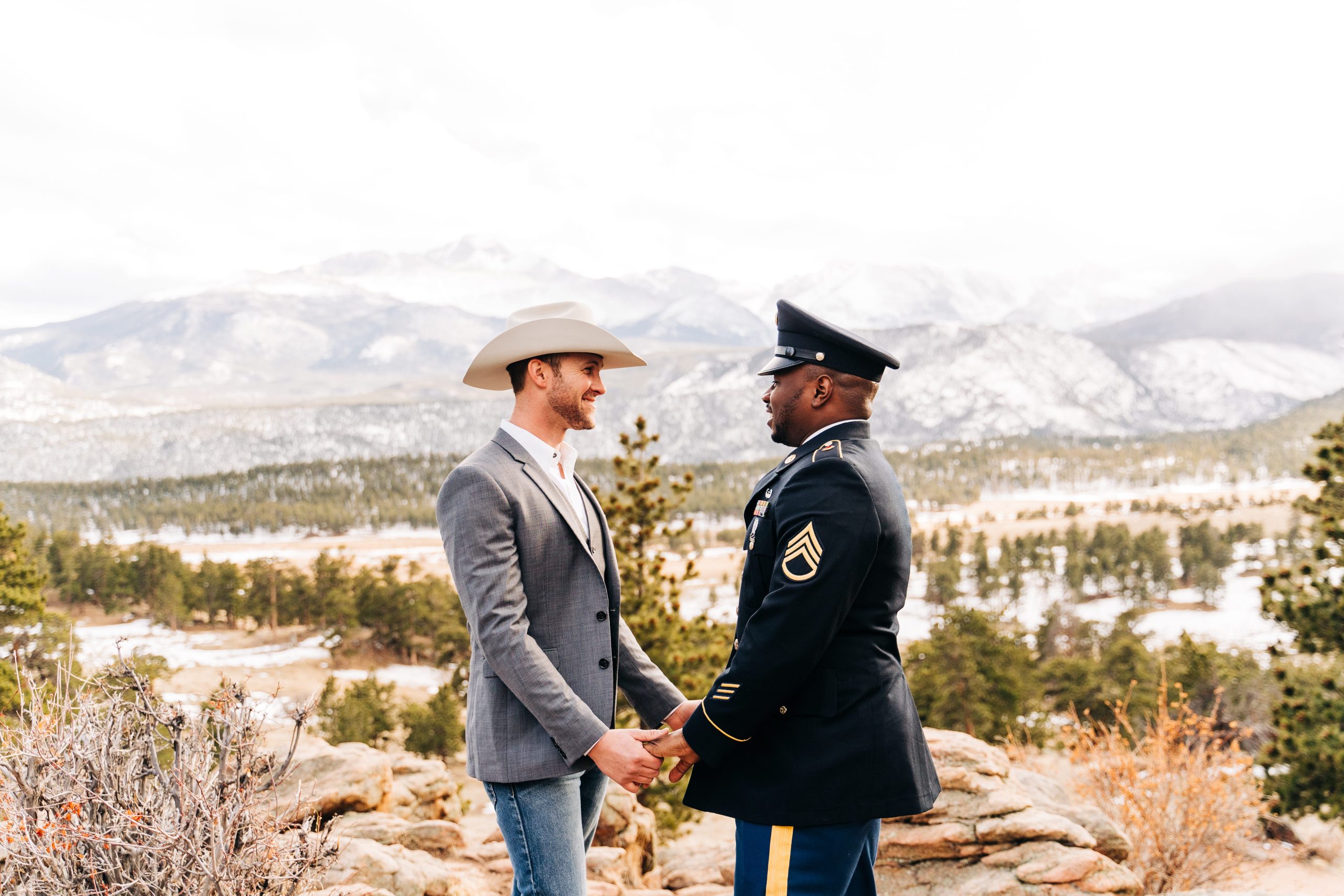 Two grooms hold hands during a mountain elopement in Colorado, standing on a rocky overlook with pine trees and snow-dusted mountains in the background, one wearing a cowboy hat and the other in military dress uniform.