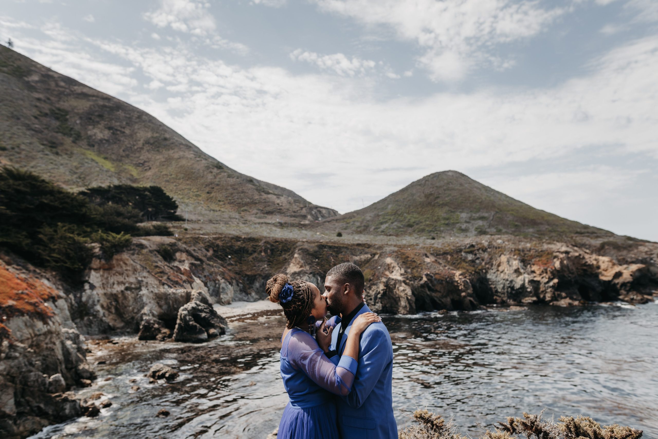 Couple share a kiss during a Big Sur elopement on a rocky coastline, wearing blue wedding outfits while standing above the Pacific Ocean with rugged cliffs in the background.
