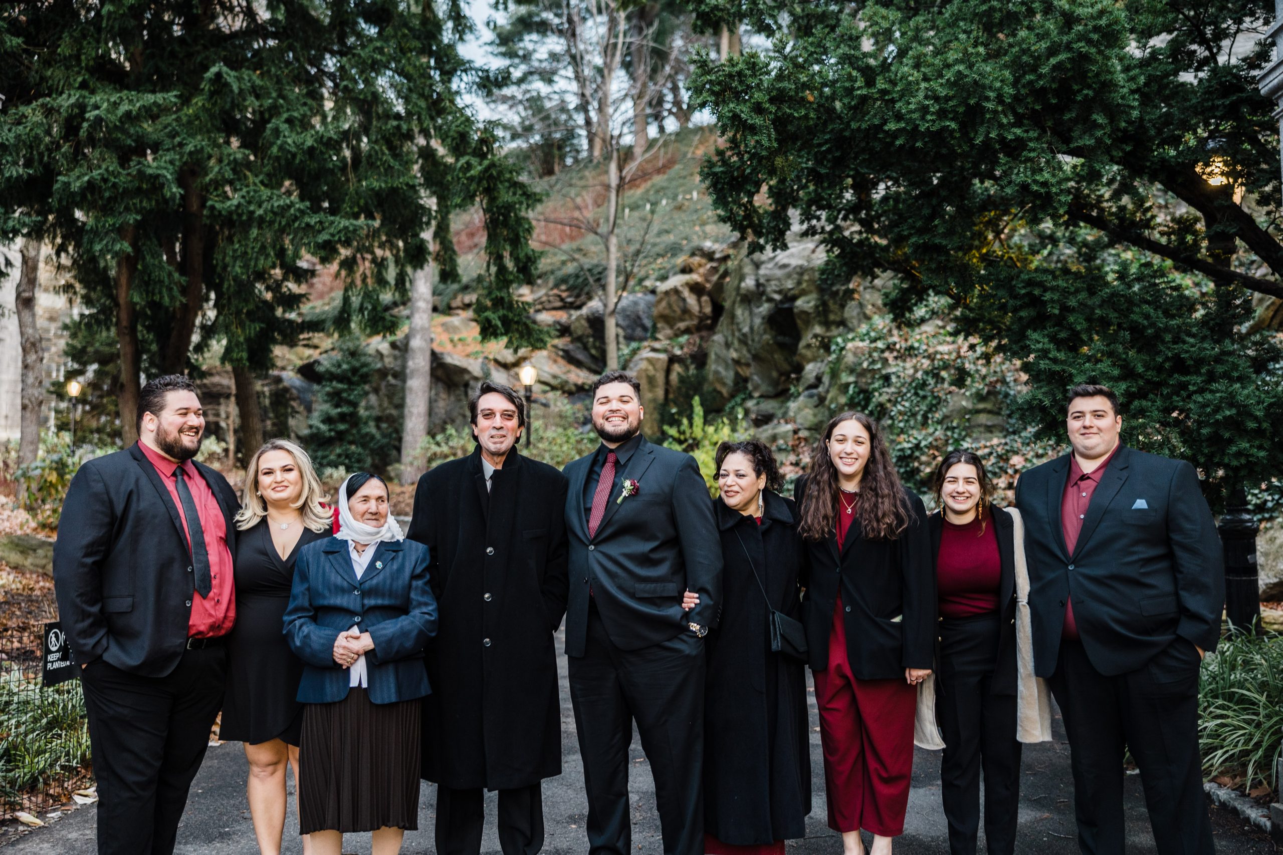 A large group of wedding guests and the groom posing outdoors in a wooded area, wearing winter wedding guest outfits in shades of burgundy and black, including long coats and formal suits.