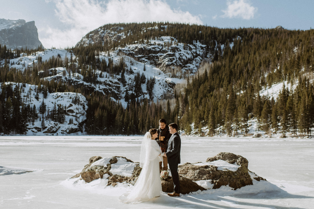 A wide shot of a wedding ceremony taking place on a frozen, snow-covered lake surrounded by a dense pine forest and snow-dusted mountains. The bride, wearing a white gown and a white fur stole, stands facing the groom in a dark suit on a small rocky outcrop. An officiant stands between them as they hold hands. The scene is bright and airy under a clear blue sky.