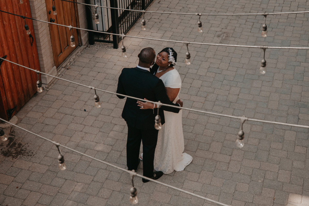 A high-angle, overhead shot of a couple embracing in a cobblestone courtyard at the Colvin House in Chicago after their winter elopement. The bride, wearing a white short-sleeved lace gown and a floral hairpiece, looks up lovingly at the groom. The groom, in a dark black suit, has his back to the camera with his hands on her waist. Several rows of Edison-style string lights crisscross over the courtyard, creating a warm, industrial-chic atmosphere.