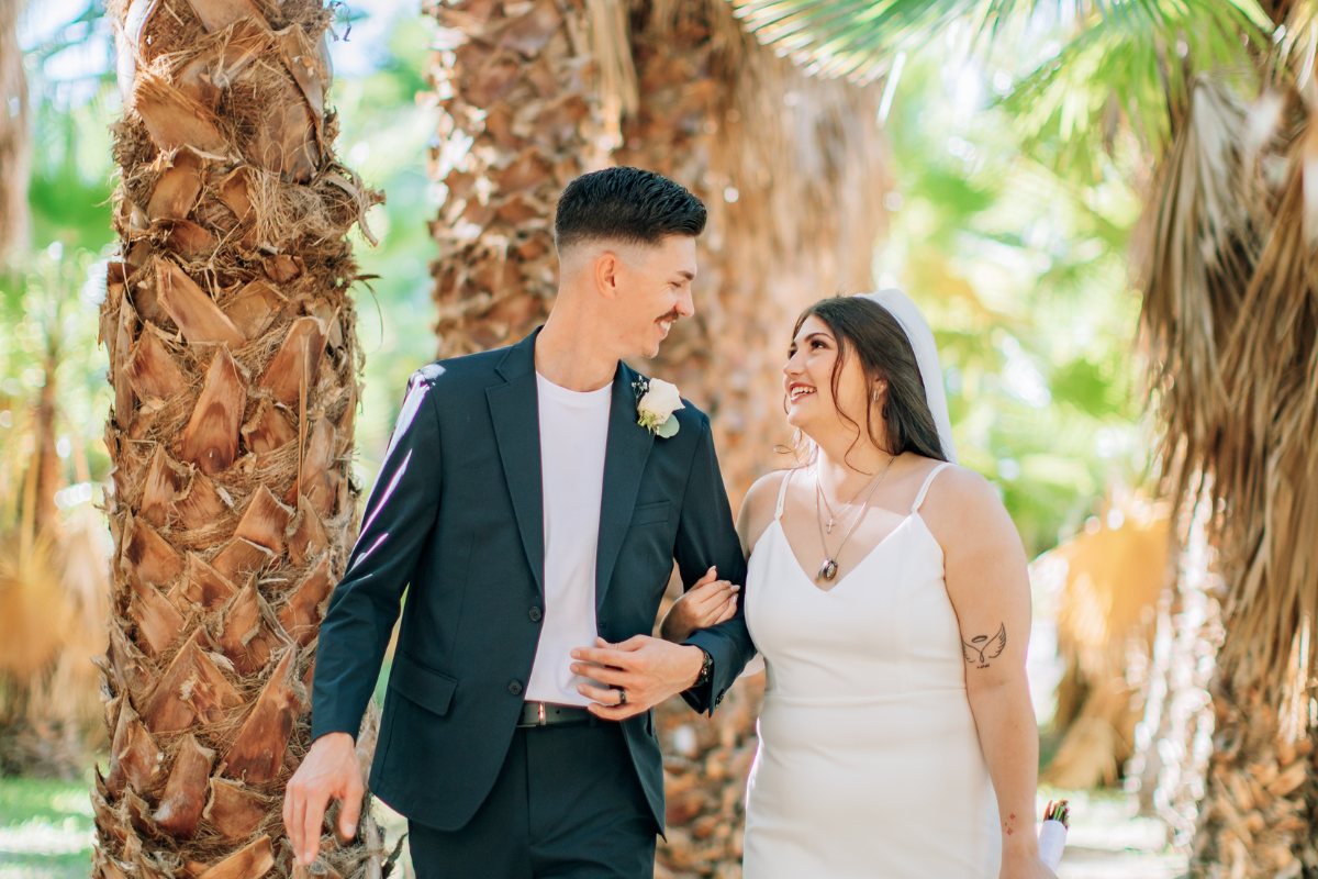 A bright, sun-drenched photo of a wedding couple walking through a lush palm grove at Greengale Farms in Las Vegas. The bride, in a white spaghetti-strap dress and veil, smiles warmly at the groom as she links arms with him. The groom, wearing a dark navy suit over a white t-shirt, looks back at her with a grin. Large, textured palm tree trunks and bright green fronds create a soft, tropical-style background.