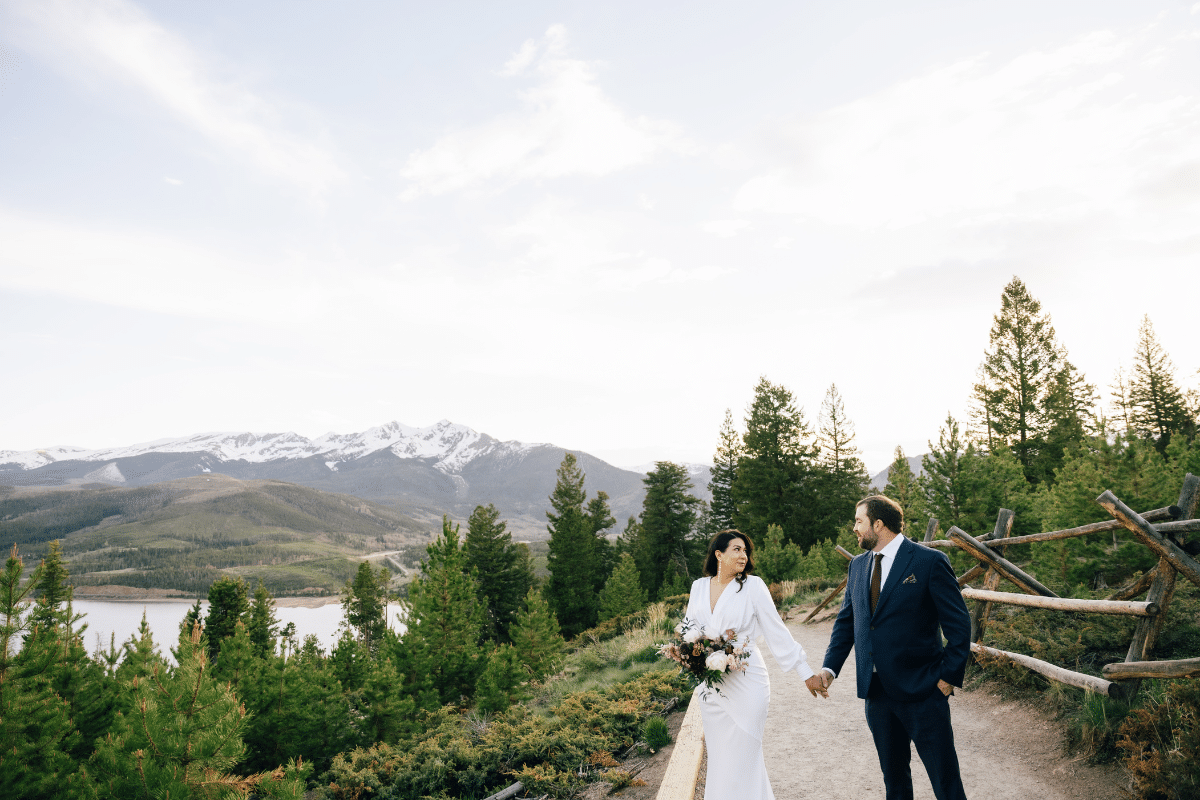 A bride in a long-sleeved white wedding dress and a groom in a navy blue suit holding hands on a dirt path during their mountain elopement wedding. They are looking at each other with snow-capped mountains, a lake, and evergreen trees in the background under a bright sky.