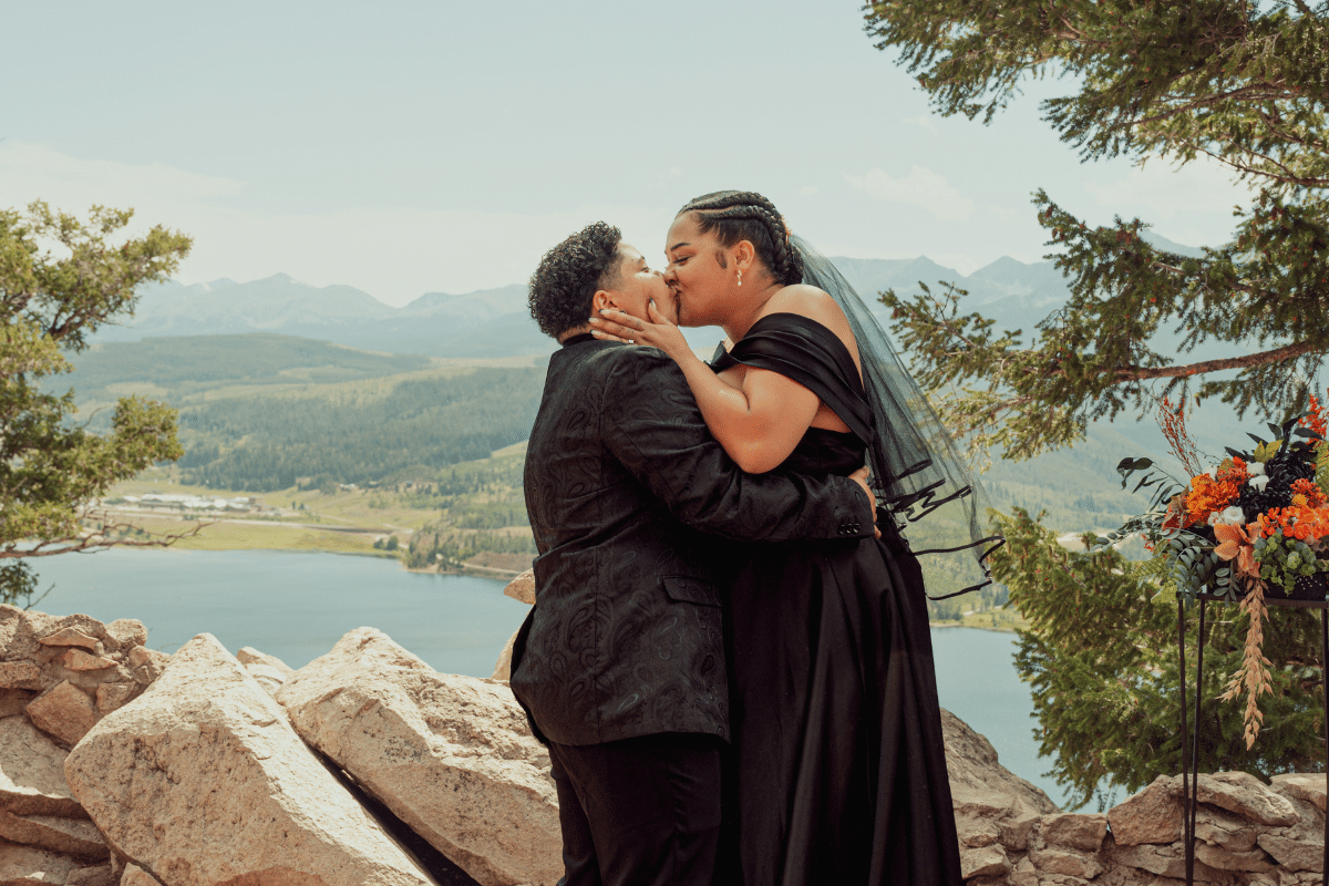 A same sex couple in stylish all-black attire sharing a passionate kiss on a rocky cliffside. One partner wears an off-the-shoulder black gown with a black veil, and the other wears a black patterned suit. They are framed by a scenic mountain range and a blue lake in the valley below.