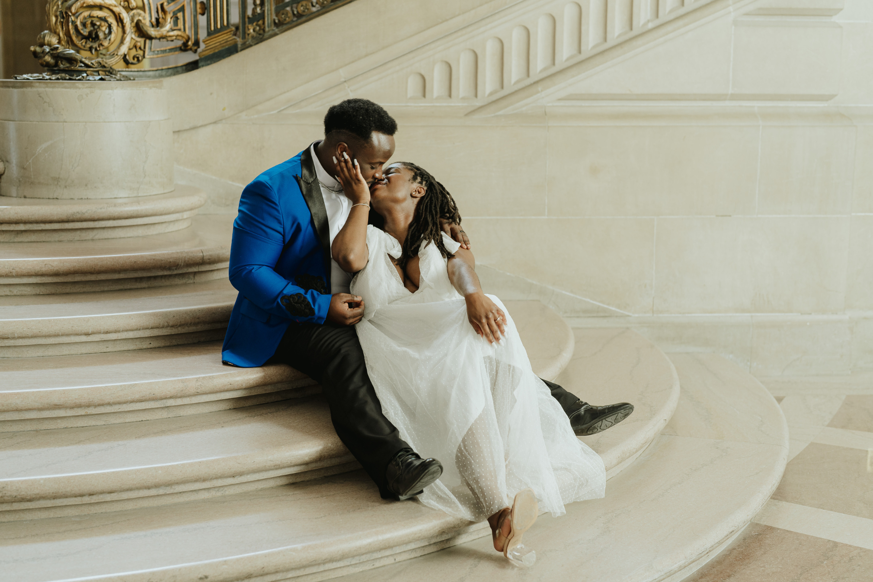 A newlywed couple sharing an intimate kiss while sitting on the grand limestone staircase of San Francisco City Hall. The groom wears a vibrant royal blue suit jacket with black lapels, and the bride wears a white polka-dot tulle wedding dress.