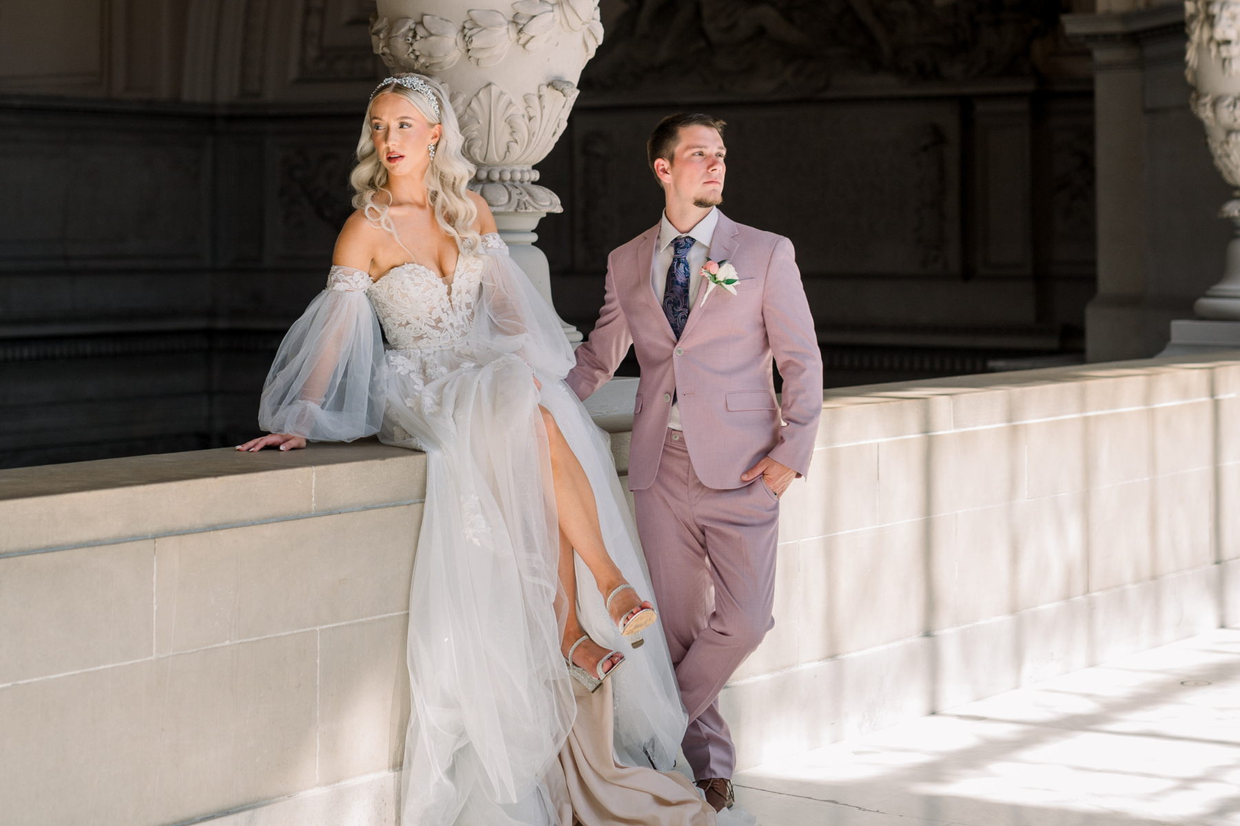 Option 1 (Descriptive):A stylish bride and groom posing against a limestone balcony at San Francisco City Hall. The bride, with long blonde waves and a jeweled headband, sits on the ledge wearing an off-the-shoulder gown with sheer puff sleeves. The groom stands beside her in a mauve-pink suit with a floral boutonniere.