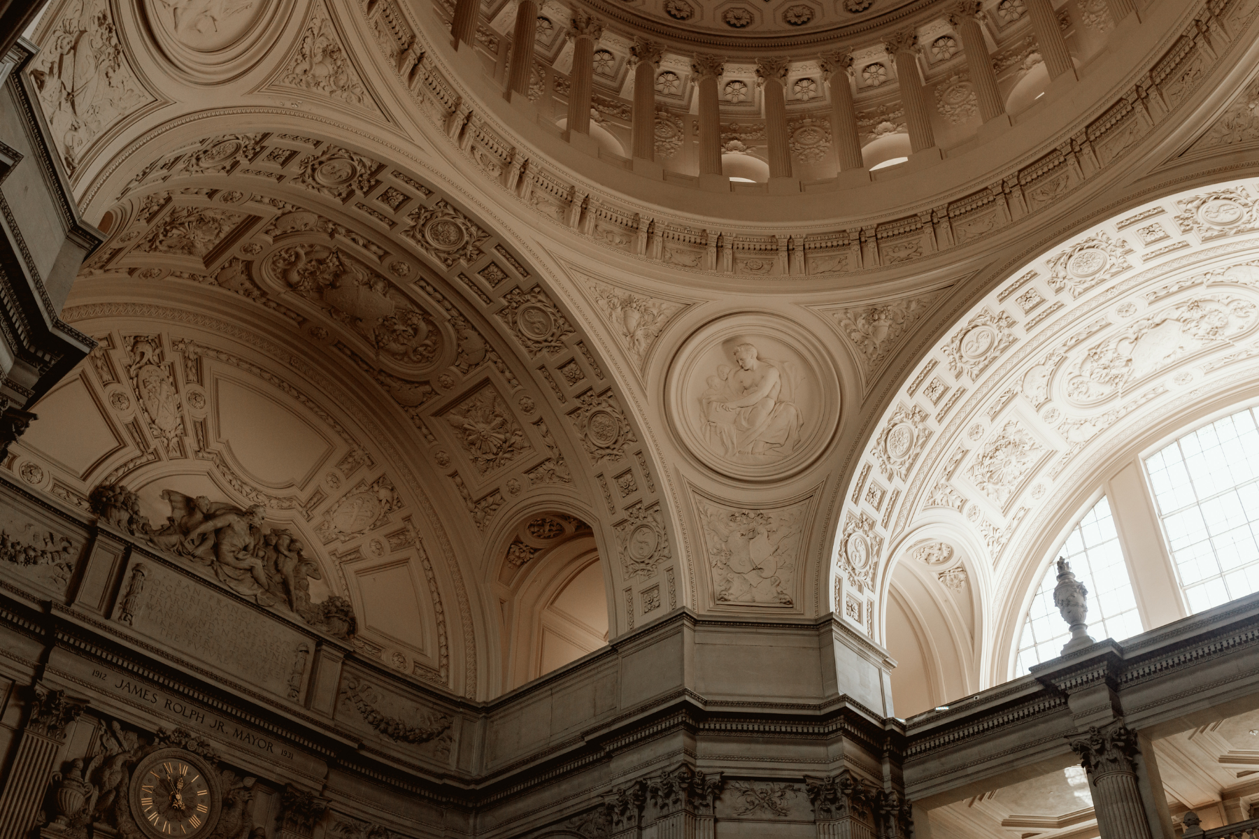 A low-angle architectural shot of the interior dome and ceiling of San Francisco City Hall. The image showcases intricate Beaux-Arts details, including ornate limestone carvings, relief sculptures, grand arches, and a view up into the columned rotunda. A historic gold clock is visible in the lower-left corner.