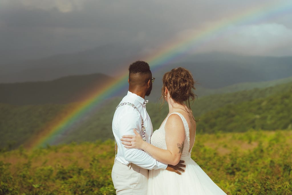 Couple holding each other looking out at North Carolina mountains as a rainbow emerges from the sky